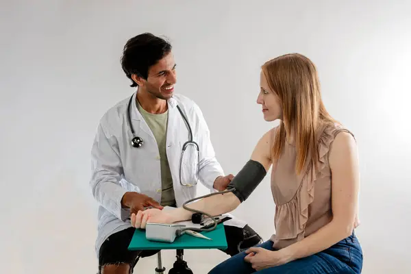 Asian doctor, from India, dressed in a white coat, measuring blood pressure on a redheaded European woman.
