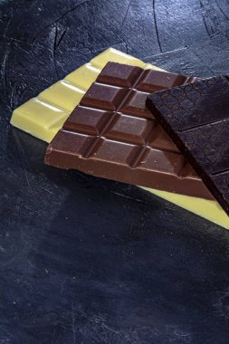 Koyu arkaplanda üç çikolata. Siyah çikolata tabletleri, beyaz ve sütlü, eskimiş görünümün koyu arka planında.