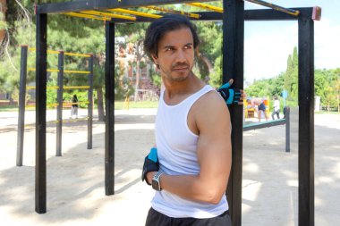 Mavi bileklikli bir adam metal bir yapının önünde duruyor. Beyaz bir atlet ve siyah bir şort giyiyor.