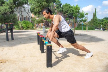 Bir adam park bankında geriniyor. Bank kırmızı ve siyah. Adam beyaz gömlek ve siyah şort giyiyor.