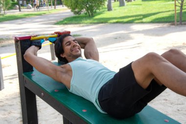 Kamu parkındaki bir bankta bacak egzersizi yapan bir adam. Yeşil tişört ve siyah pantolon giyiyor..