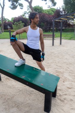 Beyaz atletli ve siyah şortlu bir adam parkta bir bankta oturuyor. Sağ elinde mavi ve gri bir eldiven var.