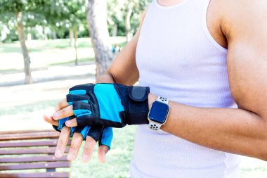 Beyaz atletli ve siyah şortlu bir adam mavi bileklik takıyor. Bir parkta duruyor ve bir şeye odaklanmış durumda.