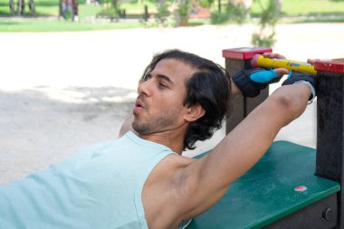 Bir adam kolları açık bir bankta uzanıyor. Yeşil gömlek ve siyah eldiven giyiyor.