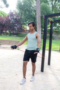 Arka planda bank olan bir adam parkta egzersiz yapıyor. Bir direniş bandı tutuyor ve antrenmanına odaklanmış durumda.