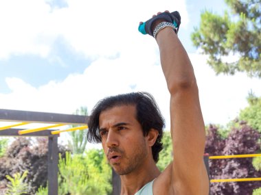 Bir adam, mavi ve yeşil atletli ve siyah şortlu bir parkta egzersiz yapıyor. Bir ip tutuyor ve bir şey kaldırıyor.