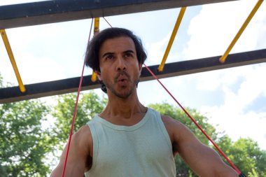 Bir adam direniş grubuyla egzersiz yapıyor. Yeşil bir atlet giyiyor ve kollarını kavuşturuyor.