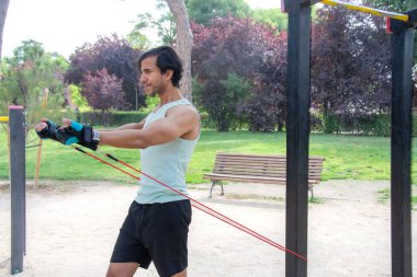 Arka planda bank olan bir adam parkta egzersiz yapıyor. Bir direniş bandı tutuyor ve antrenmanına odaklanmış durumda.