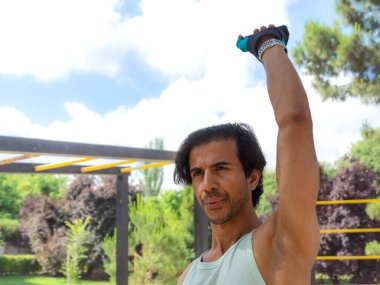 Bir adam, mavi ve yeşil atletli ve siyah şortlu bir parkta egzersiz yapıyor. Bir ip tutuyor ve bir şey kaldırıyor.