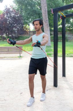 Arka planda bank olan bir adam parkta egzersiz yapıyor. Bir direniş bandı tutuyor ve antrenmanına odaklanmış durumda.