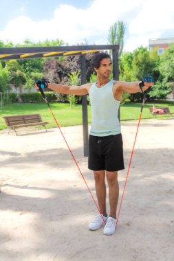 Bir adam kırmızı ve mavi bir iple parkta egzersiz yapıyor. Yeşil bir atlet ve siyah bir şort giyiyor.