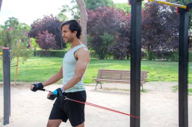 Arka planda bank olan bir adam parkta egzersiz yapıyor. Bir direniş bandı tutuyor ve antrenmanına odaklanmış durumda.