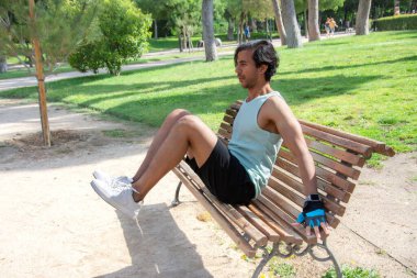 Halka açık bir parkta bankta bacak egzersizi yapan bir adam. Yeşil bir atlet ve siyah pantolon giyiyor..