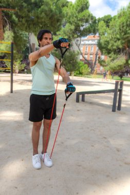 Bir adam kırmızı ve mavi bir iple parkta egzersiz yapıyor. Yeşil bir atlet ve siyah bir şort giyiyor.