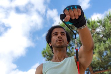 Bir adam kırmızı ve siyah direniş bandı tutuyor. Yeşil bir atlet giyiyor. Adam bir parkta ve gökyüzü mavi.