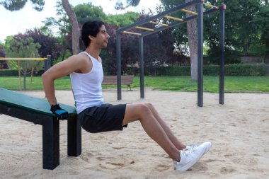 Beyaz kolsuz tişörtlü bir adam parkta bir bankta oturuyor. Kürsü, sarı direkli metal bir yapının yanında.