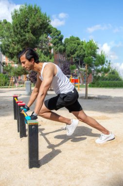 Bir adam park bankında geriniyor. Bank kırmızı ve siyah. Adam beyaz gömlek ve siyah şort giyiyor.