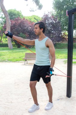 Arka planda bank olan bir adam parkta egzersiz yapıyor. Bir direniş bandı tutuyor ve antrenmanına odaklanmış durumda.