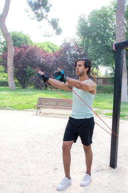 Arka planda bank olan bir adam parkta egzersiz yapıyor. Bir direniş bandı tutuyor ve antrenmanına odaklanmış durumda.