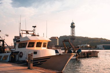 Güneş batarken Barcelona balıkçı limanına demirlemiş balıkçı tekneleri liman faaliyetlerini ve ikonik yapıları vurguluyor. Görüntü sakin bir deniz sahnesini gösteriyor..