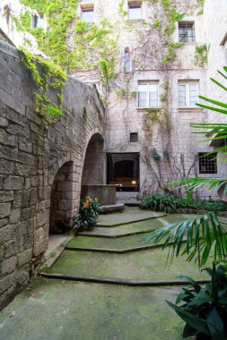 Gironas El Call mahallesindeki taş avluda kemerli kapılar, sarmaşık kaplı duvarlar ve yemyeşil alanlar var. Basamaklar, saksı bitkilerinin bulunduğu merkezi bir alana çıkıyor. Sakin ve tarihi bir ortam yaratıyor.