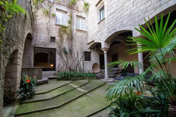 Gironas El Call mahallesindeki taş avluda kemerli kapılar, sarmaşık kaplı duvarlar ve yemyeşil alanlar var. Basamaklar, saksı bitkilerinin bulunduğu merkezi bir alana çıkıyor. Sakin ve tarihi bir ortam yaratıyor.