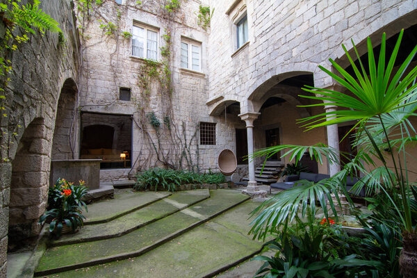 Каменный двор в районе Gironas El Call, с арочными дверями, покрытыми плющом стенами и пышной зеленью. Шаги привести к центральной области с горшком растений, создавая спокойный, исторический