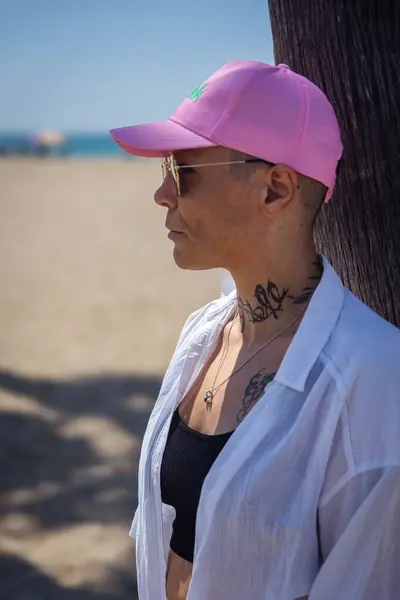 stock image Side profile of a woman with cancer wearing a pink cap and sunglasses, standing near a tree on a sunny beach.