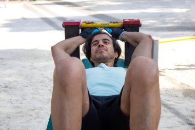 Kamu parkındaki bir bankta bacak egzersizi yapan bir adam. Yeşil tişört ve siyah pantolon giyiyor..