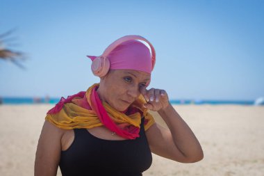 Orta yaşlı kanserden kurtulan, pembe kulaklıklar takan ve başörtüsü takan, sahildeki bir gözyaşını silen, incinebilirliği ve duygusal gücü temsil eden biri..