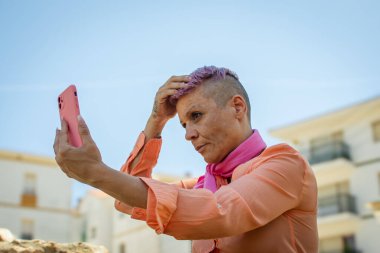 Kafası tıraşlı bir kanserden kurtulan birinin portresi açık havada dış görünüşünü kontrol ediyor. Parlak gökyüzü arkaplanı onun odaklanma ve konsantrasyonunu vurguluyor.