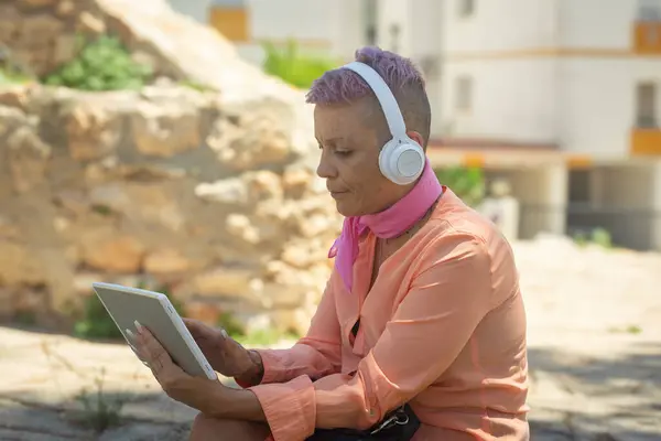 stock image Portrait of a cancer survivor with a shaved head, wearing headphones and using a tablet while sitting outdoors. The serene natural background adds to the focus and concentration on the task.