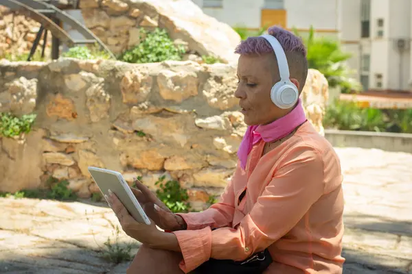 stock image Cancer survivor with a shaved head, wearing headphones and enjoying media on a tablet while sitting outdoors. The tranquil outdoor setting complements her relaxed state.