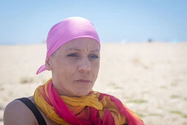 stock image Middle-aged cancer survivor in a pink headscarf, lost in thought by the seaside, reflecting on her journey