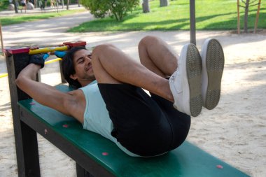 Kamu parkındaki bir bankta bacak egzersizi yapan bir adam. Yeşil tişört ve siyah pantolon giyiyor..