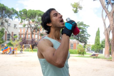 Eldiven takmış bir adam şişeden su içiyor. Sahne ağaçlı ve banklı bir parkta geçiyor. Adam antremanlarına ya da açık hava aktivitelerine ara veriyor.