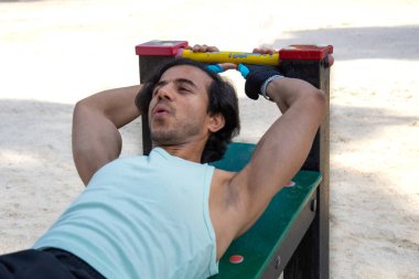 Kamu parkındaki bir bankta bacak egzersizi yapan bir adam. Yeşil tişört ve siyah pantolon giyiyor..