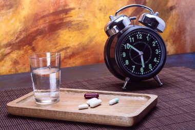 Assorted pills on a wooden tray with a glass of water and a vintage alarm clock on a table, bright background. clipart