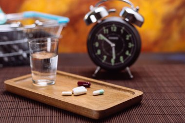 Assorted medication placed on a wooden tray with a glass of water, a vintage alarm clock, and a shopping basket filled with medicines. clipart