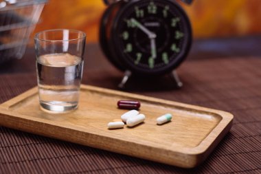 Close-up of assorted pills on a wooden tray with a glass of water, a vintage alarm clock, and a shopping basket, vibrant background. clipart