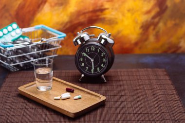 Assorted pills on a wooden tray with a glass of water, a vintage alarm clock, and a shopping basket filled with medicine, vibrant background. clipart