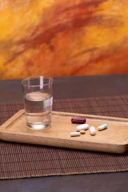 A glass of water and various pills on a wooden tray, representing a daily health routine of medication and hydration. clipart