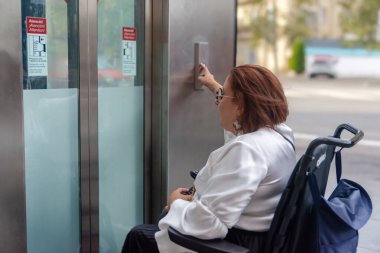 Elektrikli sandalyedeki kişi asansör düğmesine basarak erişilebilirlik özelliklerini gösterir..