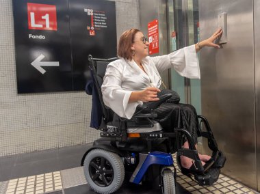 Kadın tekerlekli sandalye kullanıcısı metro istasyonundaki asansörü aramak için düğmeye basıyor ve erişilebilir tasarımı gösteriyor..