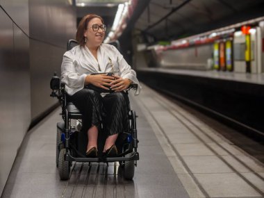 Kadın tekerlekli sandalye kullanıcısı ulaşılabilir bir platformda metro trenini bekliyor. Bu da hareket kabiliyetini gösteriyor..