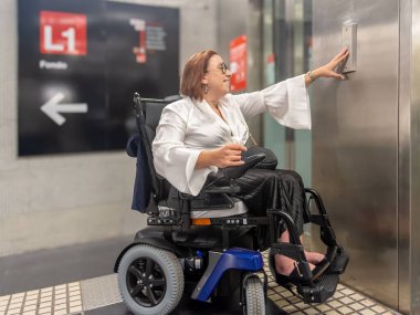 Woman using an electric wheelchair presses the elevator button at the metro station for accessible entry.