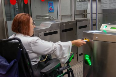 Kadın tekerlekli sandalye kullanıcısı, toplu taşıma araçlarını gösteren ulaşılabilir bir metro kapısında geçiş kartını kullanıyor..