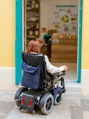 Tekerlekli sandalyedeki bir kadın, bir mağaza girişinde, erişilemez alanların zorluklarını vurgulayan bir basamakla karşılaşır..