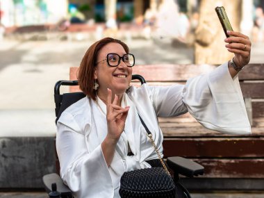 Tekerlekli sandalyede gülümseyen ve parktaki akıllı telefonuyla selfie çeken bir kadın..