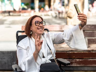 Kadın tekerlekli sandalye kullanıcısı rahat bir şehir ortamında selfie çekmekten zevk alıyor, yetki ve bağımsızlık gösteriyor.